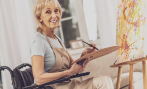 Smiling blonde woman seated at an easel holding an artists' palette and paintbrush.