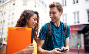 Two students walking in the city while chatting. One is holding a clipboard.