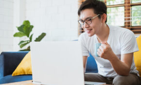 A young man sitting on a sofa looking at an open laptop on the coffee table in front of him; he is holding a mug and smiling