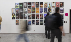 People walk by a series of framed illustrations hung in a grid pattern on a wall in an ECU campus gallery.